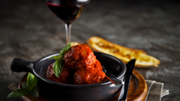 three italian meatballs in napoli sauce topped with basil in a dark bowl with glass of red wine in the background